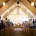 A blurred photo of the inside of a church sanctuary that is filled with people in the pews, and the pastor stands under a large cross at the altar.