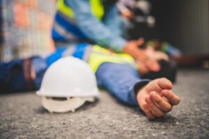 blurred image of an injured worker lying on the ground next to a hardhat with coworker checking on him