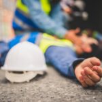 blurred image of an injured worker lying on the ground next to a hardhat with coworker checking on him