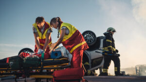 paramedics attending to a car accident victim on a stretcher with overturned vehicle behind them