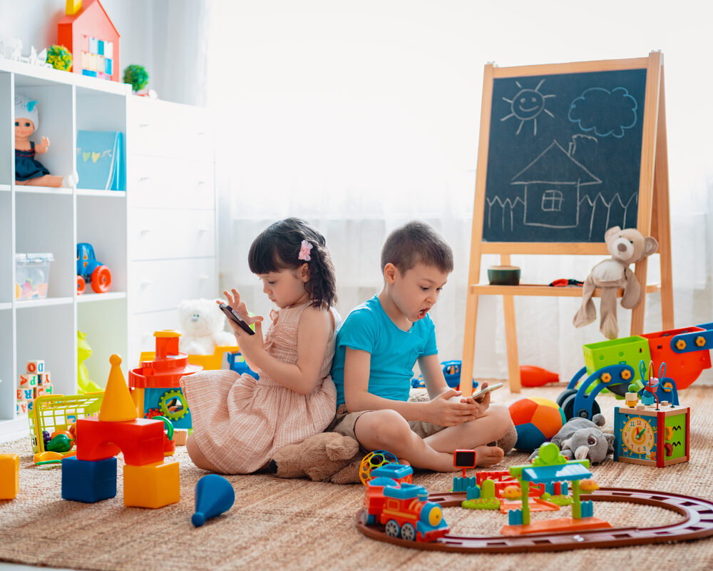 Siblings children brother and sister sit on the floor of the house in the children's play room with smartphones, detached from the scattered toys.