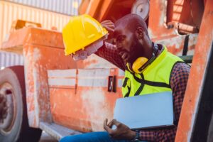 african american worker sweating while sitting on heavy machinery holding his hardhat from hot weather in summer working at port 