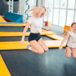 An older and younger sister bounce on a trampoline indoors