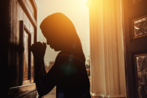 silhouette of young woman kneeling and praying in church with sunlight shining