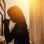 silhouette of young woman kneeling and praying in church with sunlight shining