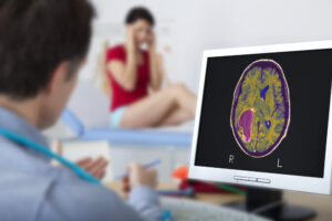 a young woman on a doctor table holding her head while male doctor looks at a brain tumor on ct scan screen