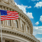 American flag flies in front of the US capitol building in Washington DC with blue cloudy sky background