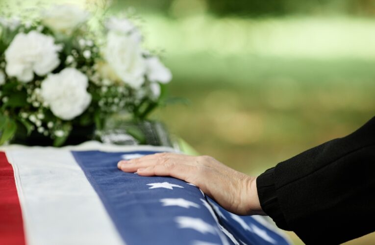 closeup of a hand on a veteran's coffin at funeral with white flowers in the background