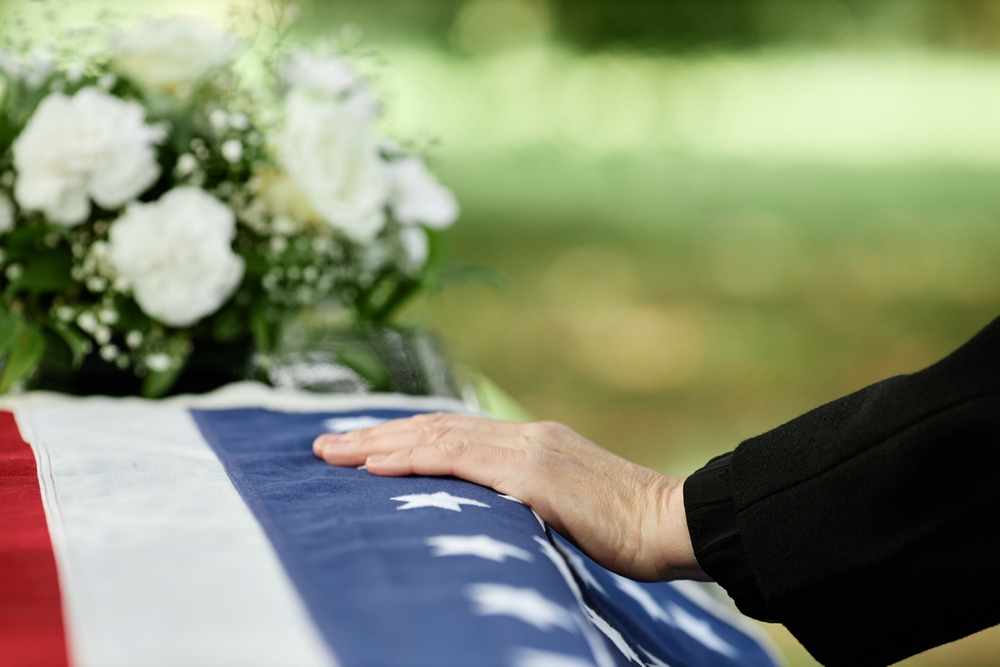 closeup of a hand on a veteran's coffin at funeral with white flowers in the background